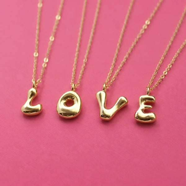 Personalised gold bubble initial necklaces spelling LOVE with chunky letter pendants on a pink background