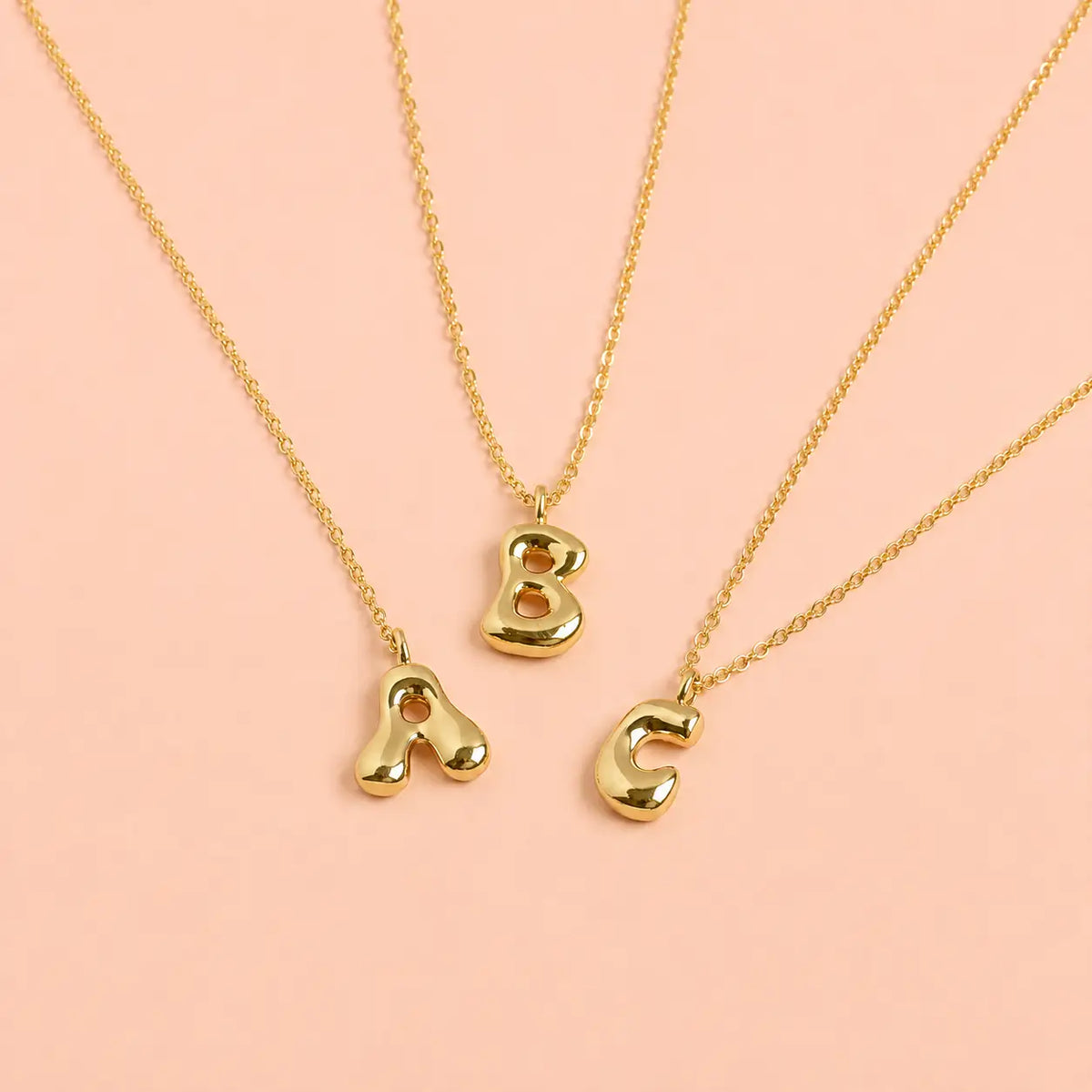 Personalised gold bubble initial necklaces with A B and C letter pendants on a pale pink background