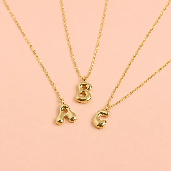 Personalised gold bubble initial necklaces with A B and C letter pendants on a pale pink background