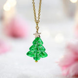 image shows a green glass Christmas tree necklace with gold star detail on a pink festive background