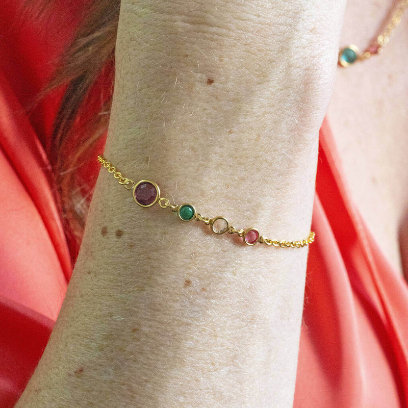 A gold mother and three children round birthstone bracelet worn on a wrist. The bigger round birthstone is Amethyst followed by three smaller round birthstones, Emerald, Crystal and Rose.