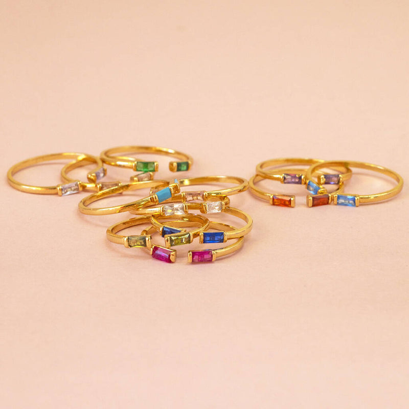 Image shows 12 Baguette Birthstone Stacking Rings for each month of the year on a pink backdrop.