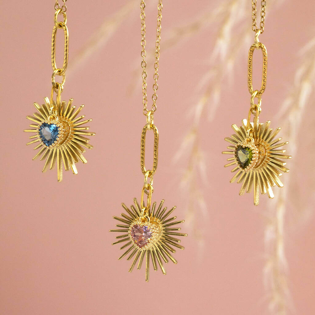 Image shows three Birthstone Heart Starburst Necklaces in front of a pink backdrop. From left to right: September Sapphire, October Rose and August Peridot.