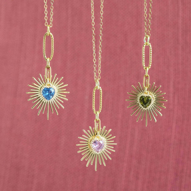 Image shows three Birthstone Heart Starburst Necklaces in front of a pink backdrop. From left to right: September Sapphire, October Rose and August Peridot.