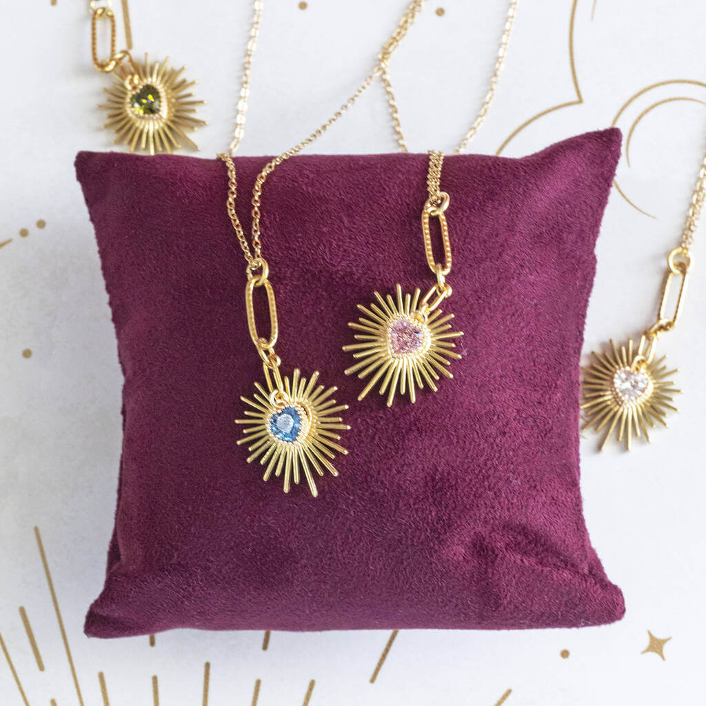 Image shows three Birthstone Heart Starburst Necklaces on a maroon jewellery pillow andwhite backdrop. From left to right: August Peridot, September Sapphire, April Crystal and October Rose.