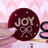 Gold beaded marron velvet JOY compact mirror set on a pink background with makeup