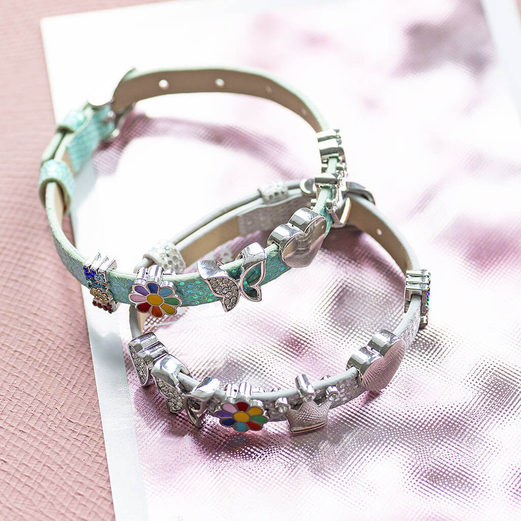 A silver and a green ,fully adjustable,faux leather buckle bracelet with sparkly heart, sparkly butterfly, enamel coloured flower, shiny crown, shiny heart and sparkly LOVE charms displayed on a sparkly pink background.