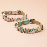 A silver and a green faux leather buckle bracelet with sparkly heart, sparkly butterfly, enamel coloured flower, shiny crown, shiny heart and sparkly LOVE charms displayed on a plain peach coloured background.