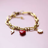Personalised gold charm bracelet with chunky chain featuring red heart, lightning bolt and clover charms on pink background