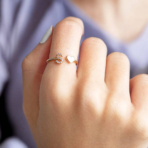 A gold plated ring with crystal encrusted initial charm and a heart charm worn on an index finger.