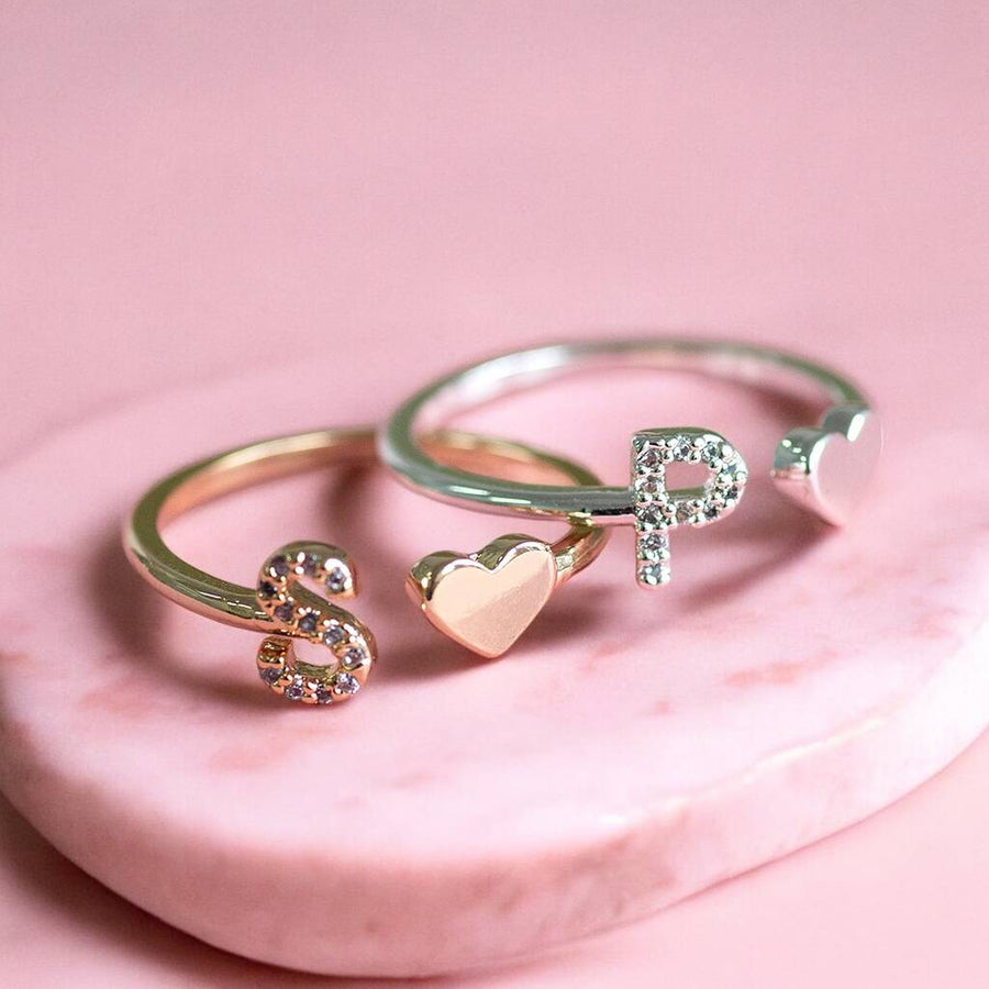 Two open band rings displayed on a pink stone surface against a pink background. One ring is gold with a small heart charm and clear stones, and the other is silver with a letter P charm and clear stones.