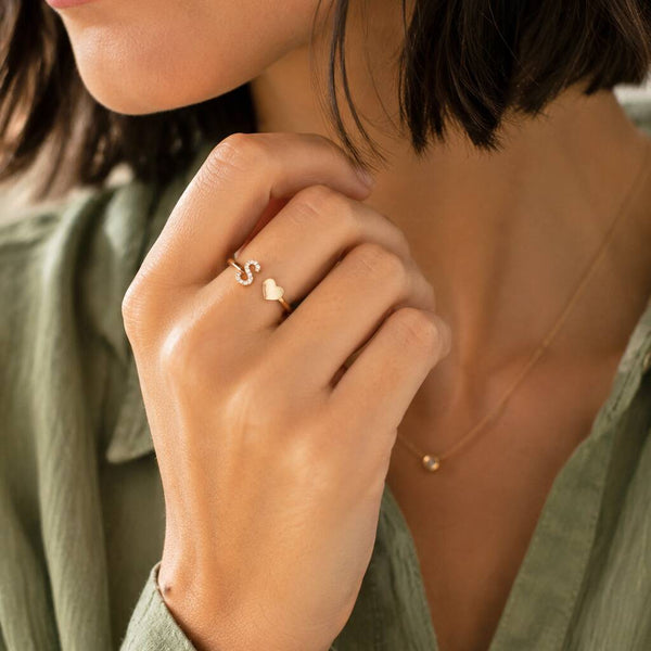 A close-up lifestyle shot of a woman wearing a delicate gold ring with a small heart and letter 'S' design on her ring finger, paired with a sage green top.