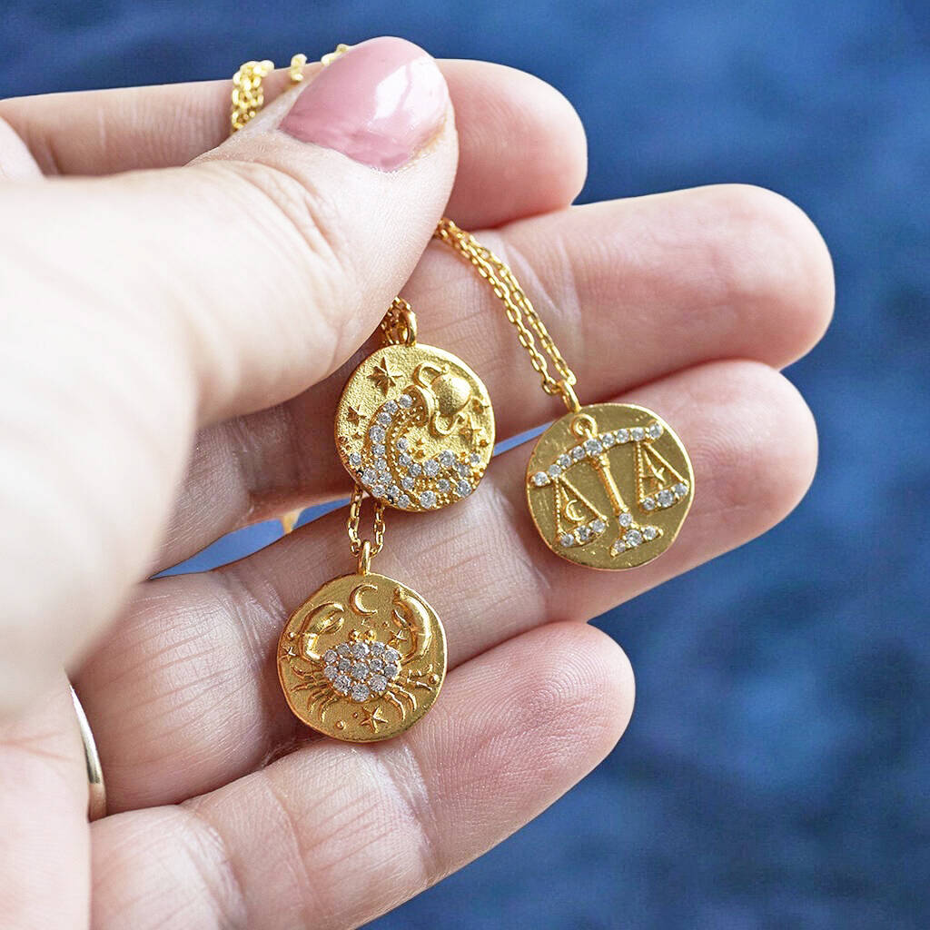 image shows three of the twelve crystal zodiac coin necklaces displayed on a woman's hand on a blue background