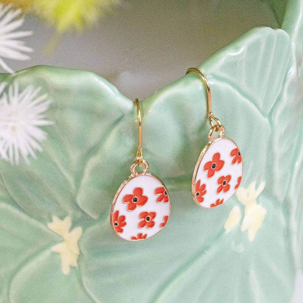 white with orange flowers enamel Easter egg charms on gold plated earring hooks display on the side of a plant pot