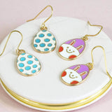 blue and white polka dot and purple white bunny face enamel Easter egg shaped charms on gold plated earring hooks displayed on a white marble coaster