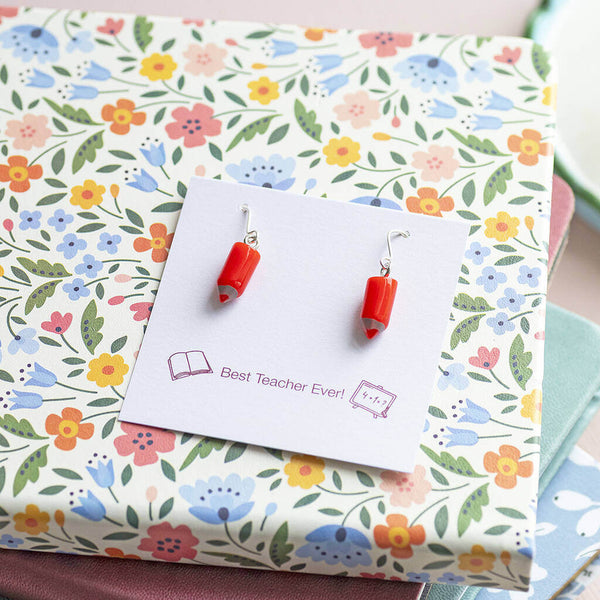 Red pencil earrings presented on a Best Teacher Ever sentiment card lying flat on top of a floral notebook