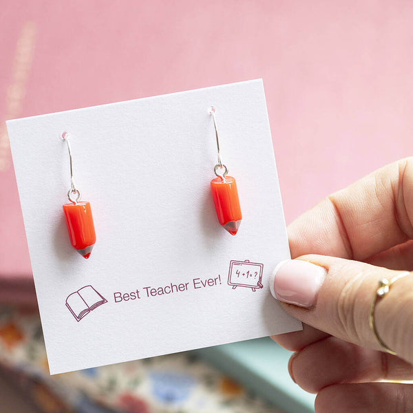 Red pencil charm drop earring presented on a Best Teacher Ever sentiment card being held against a backdrop of school jotters