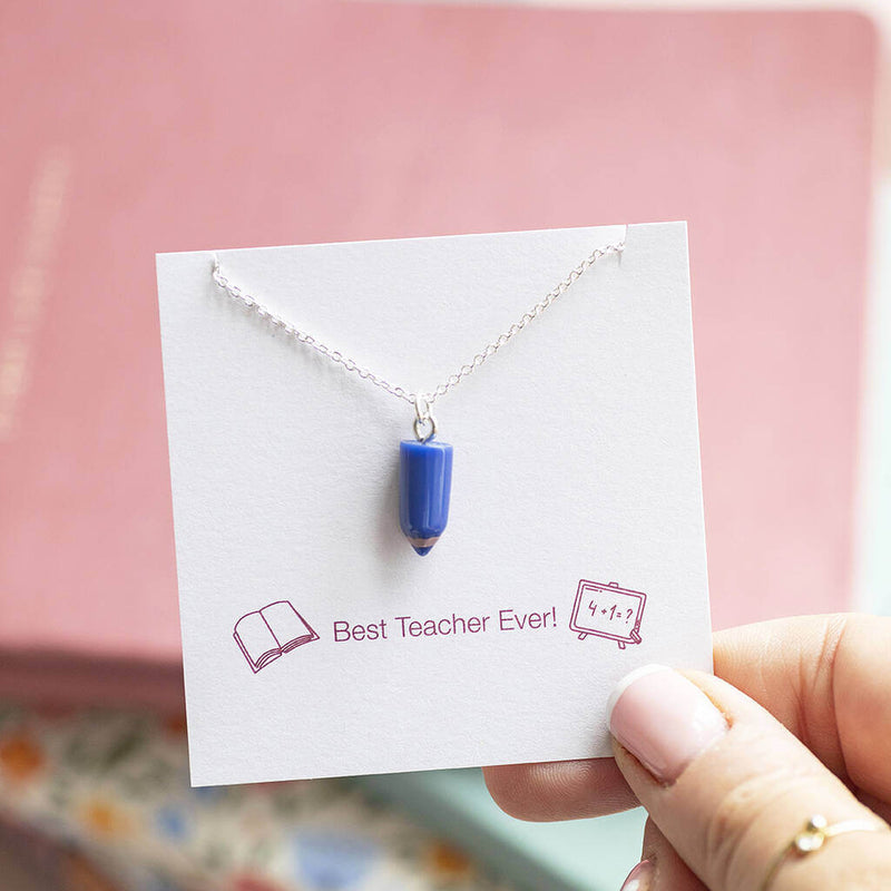 Blue pencil charm attached to a silver necklace chain displayed on a Best Teacher Ever sentiment card with pink and floral notebooks int he background.