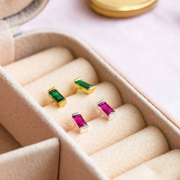 Colourful pink and green baguette birthstone studs in silver and gold tones, displayed in a soft velvet jewellery box.
