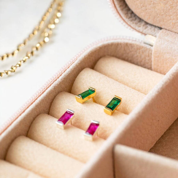 Vibrant pink and green baguette birthstone studs in silver and gold tones, styled together in a soft jewellery box.