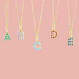 Image shows a selection of birthstone studded initial necklaces hanging in front of a pink background