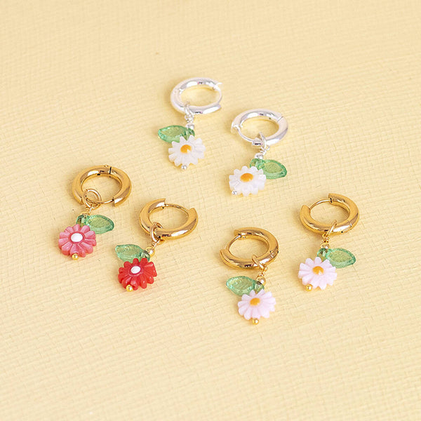 Image shows from top - silver plated white Daisy Huggie Hoop Earrings, gold plated white Daisy Huggie Hoop Earrings, pale pink gold plated Daisy Huggie Hoop Earrings and dark pink Daisy Huggie Hoop Earrings on a yellow backdrop.