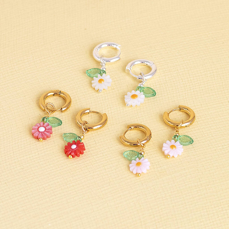 Image shows from top - silver plated white Daisy Huggie Hoop Earrings, gold plated white Daisy Huggie Hoop Earrings, pale pink gold plated Daisy Huggie Hoop Earrings and dark pink Daisy Huggie Hoop Earrings on a yellow backdrop.