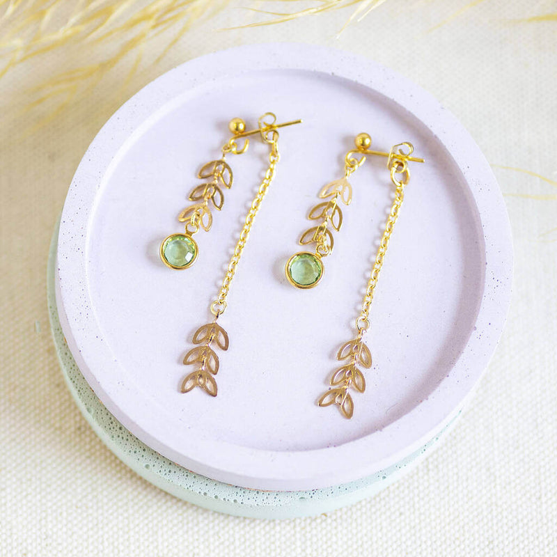 Image shows Double Drop Leaf Chain Birthstone Earrings with August Peridot birthstone on a pink backdrop.