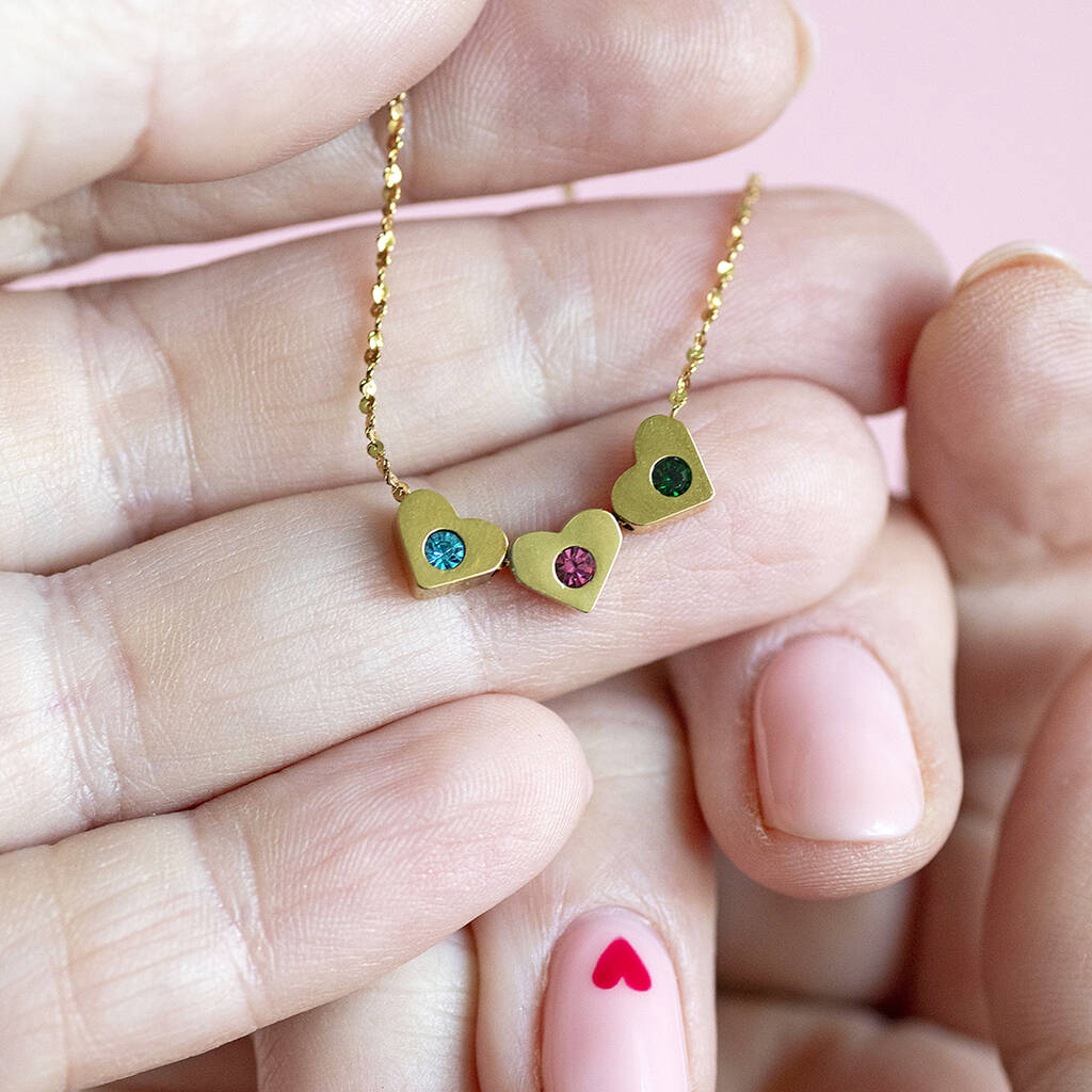 image shows hands holding a gold necklace with three hearts set with different coloured birthstones