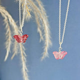 Image show red and pink glass butterfly necklaces hanging in front of a blue background