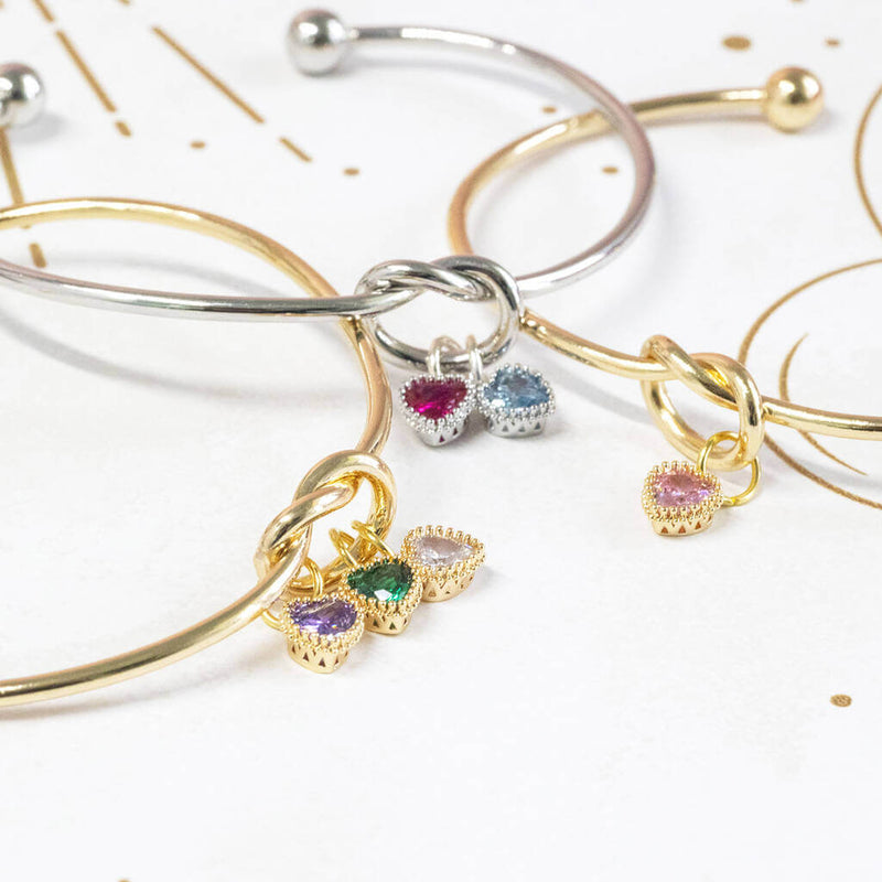 Image shows three friendship knot bangles from left to right: gold plated with three charms, silver plated with two charms and gold plated with one charm.
