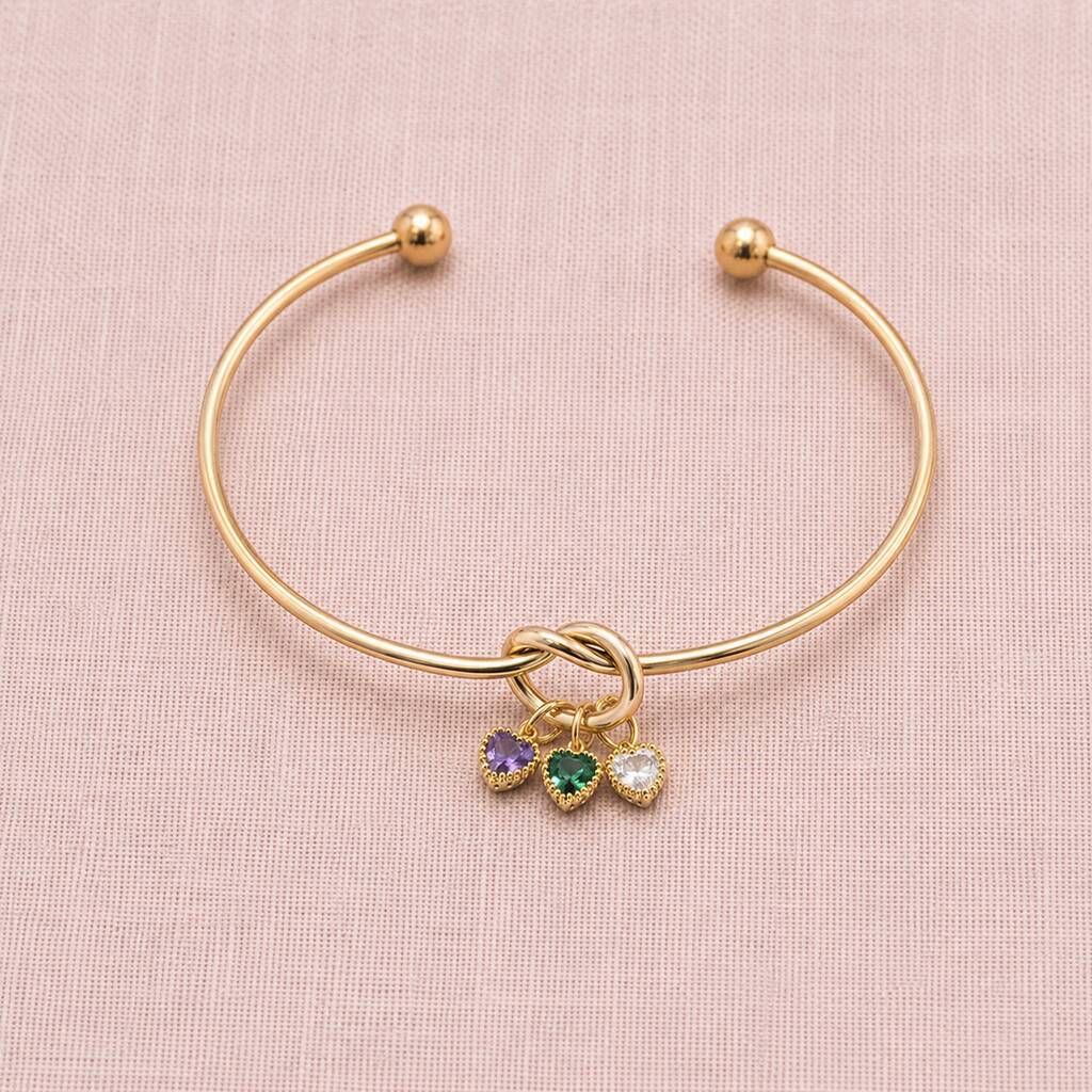 Gold plated friendship knot bangle with purple, green and clear heart birthstone charms on a soft pink backdrop