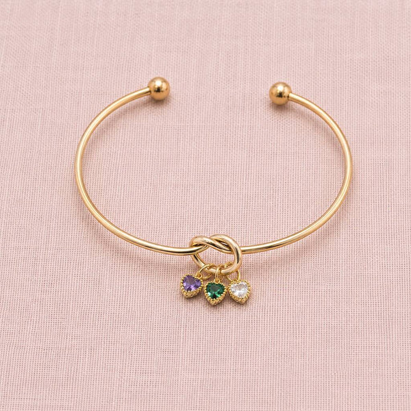 Gold plated friendship knot bangle with purple, green and clear heart birthstone charms on a soft pink backdrop