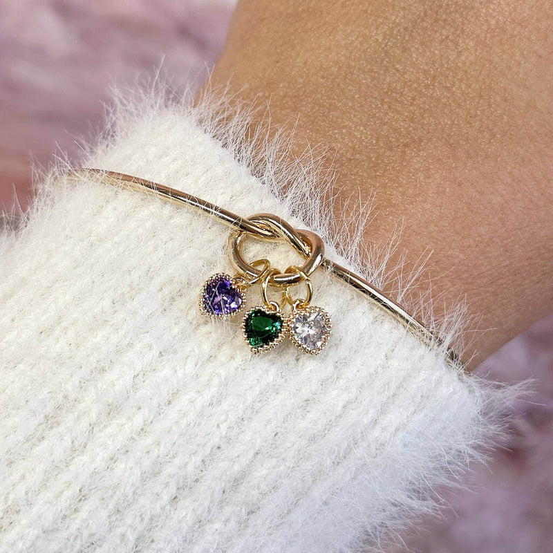 Image shows model wearing gold plated friendship knot bangle with thee birthstone hearts from left to right: February Amethyst, May Emerald and April crystal.