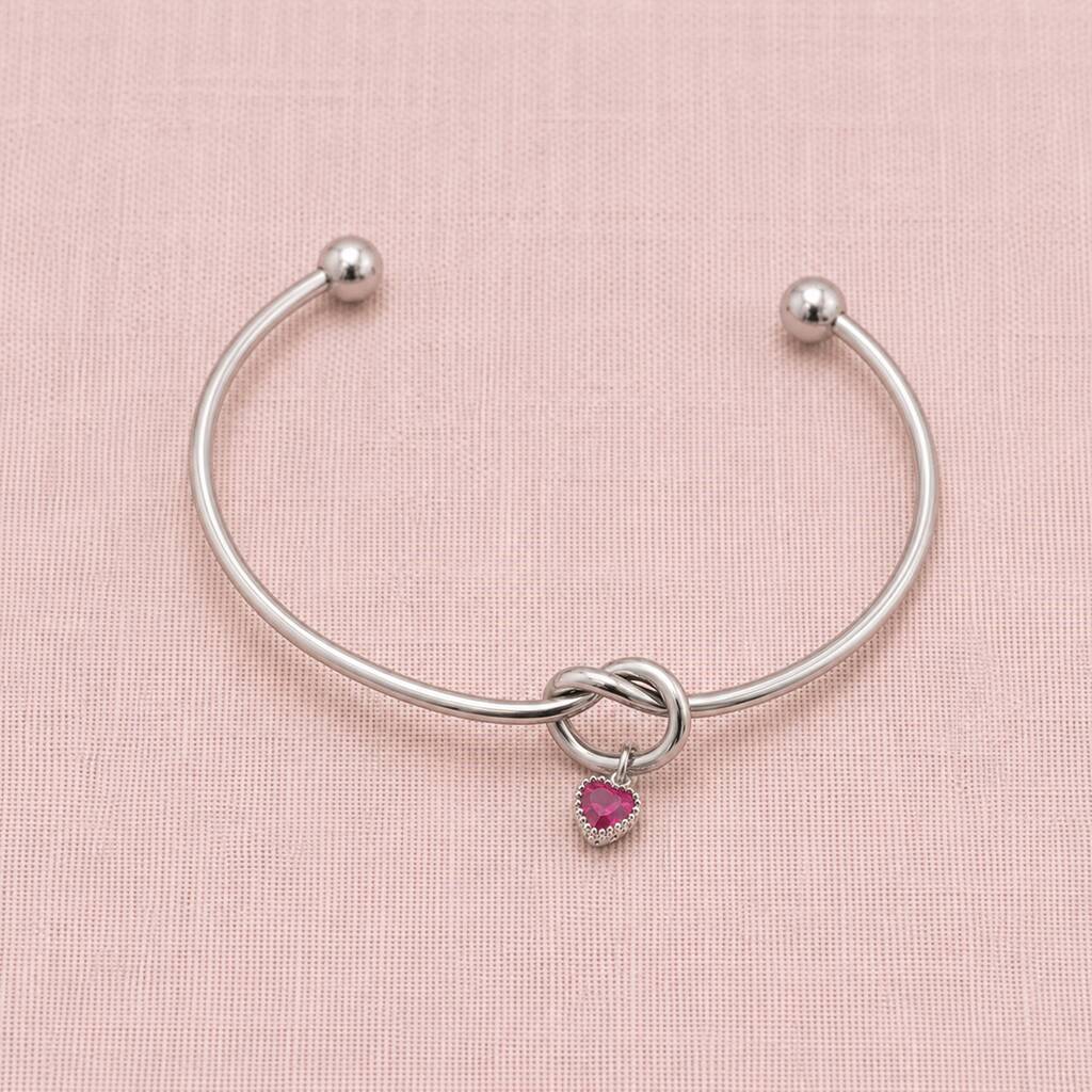 Minimal silver friendship knot bangle featuring one pink heart birthstone charm on a blush fabric background