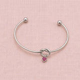 Minimal silver friendship knot bangle featuring one pink heart birthstone charm on a blush fabric background