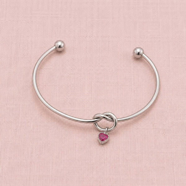 Minimal silver friendship knot bangle featuring one pink heart birthstone charm on a blush fabric background