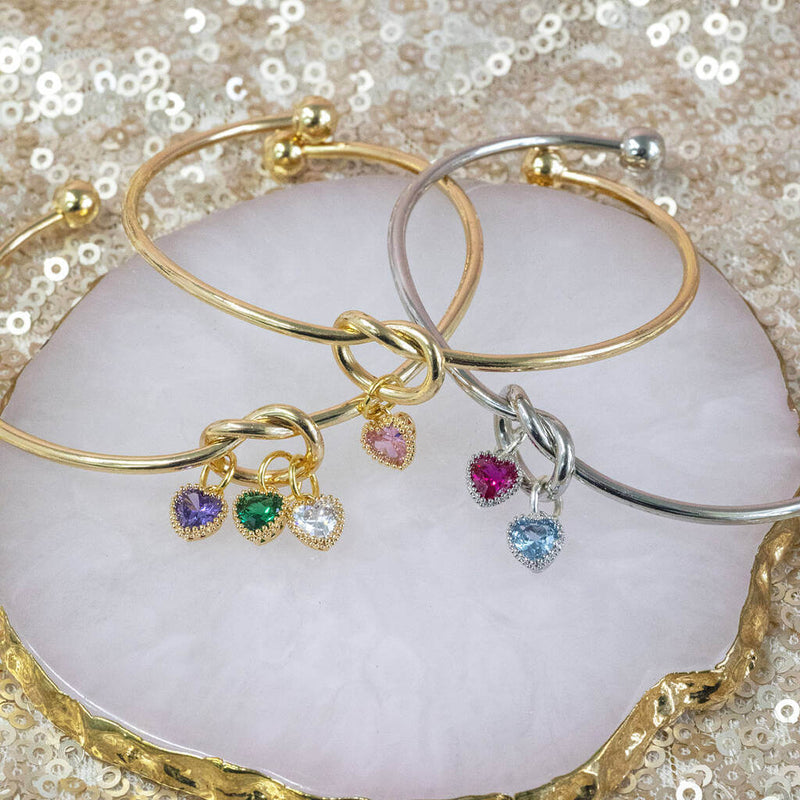 Image shows three friendship knot bangles from left to right: gold plated with three charms, gold plated with one charm and silver plated with two charms.