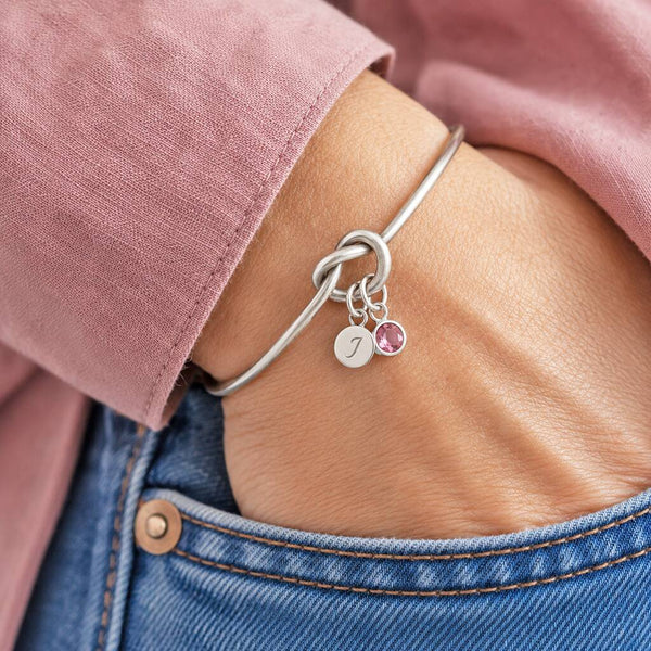 Silver friendship knot bangle with pink crystal birthstone and initial charm, perfect for stacking