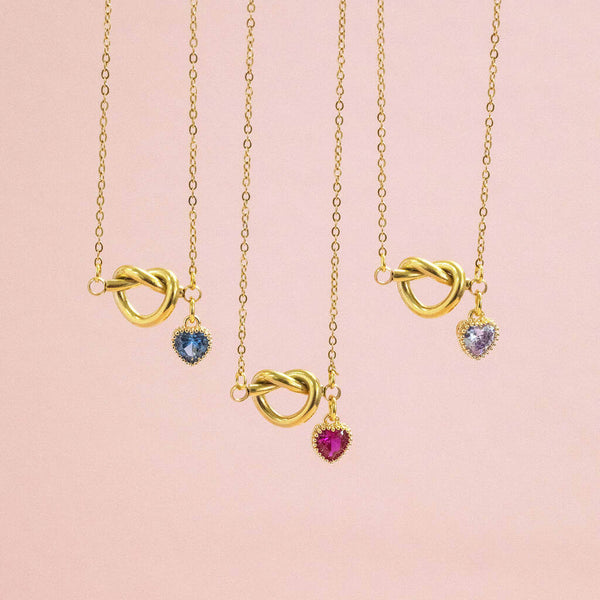 Image shows three Friendship Knot Necklace with Heart Birthstones from left to right: Sapphire, ruby and Light Amethyst in front of a pink backdrop.