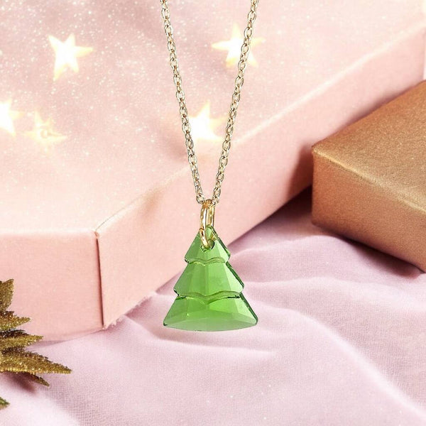 Image shows green glass Christmas tree charm suspended from a gold plated trace chain on a pink back ground with small gold stars.