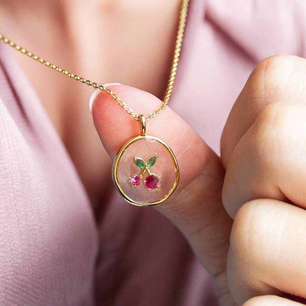 A round clear glass pendant suspended from a gold chain with a crystal cherry charm.