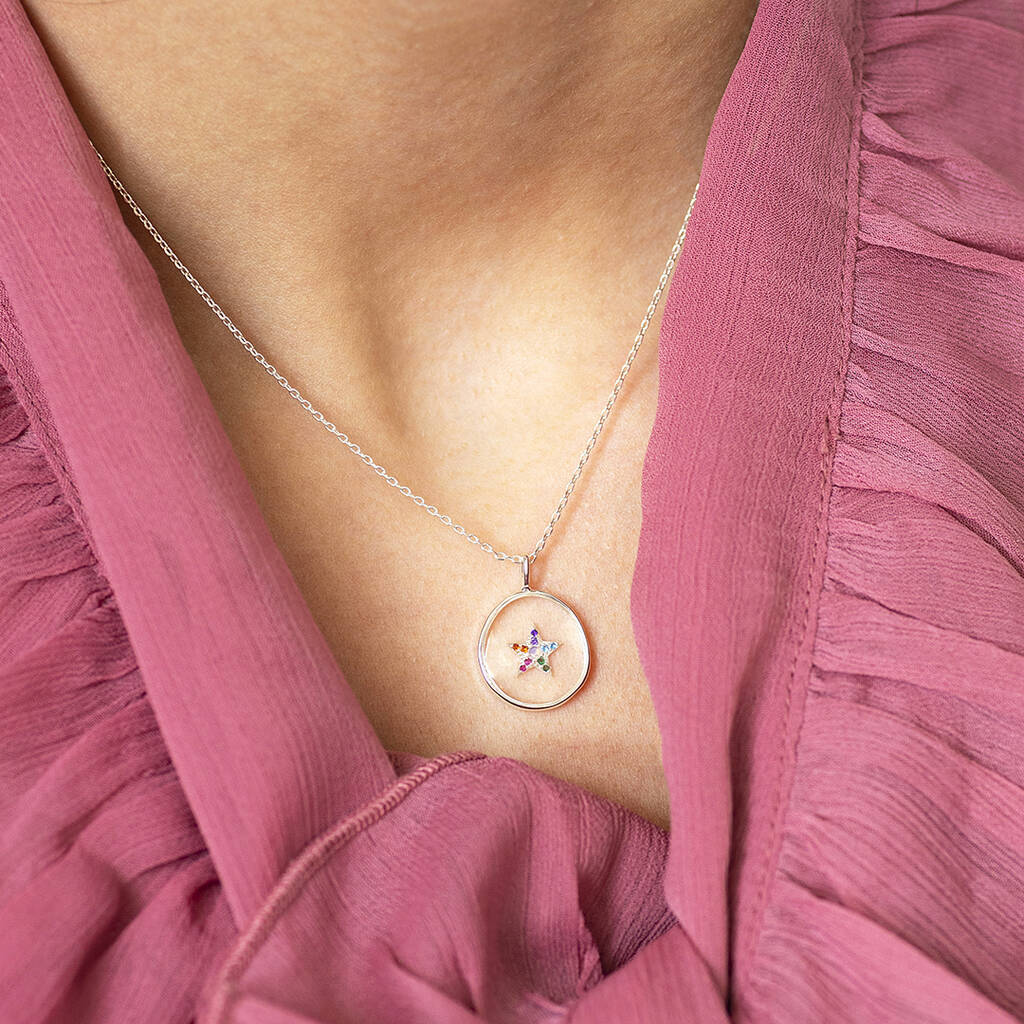 Image shows a multi coloured crystal star clear glass round pendant suspend from a silver trace chain. Worn with a pink ruffle detail top.