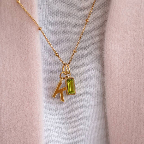 Gold plated initial necklace with green baguette birthstone charm, layered charm jewellery with colourful styling appeal