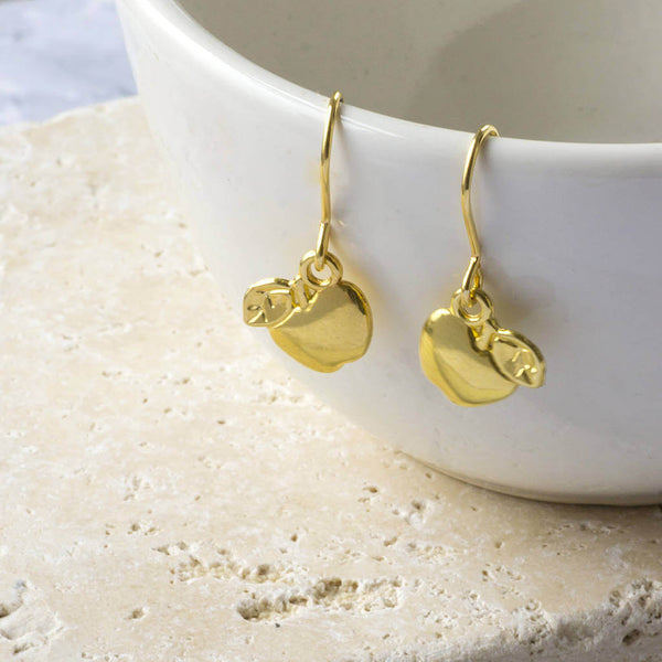 Gold apple drop earrings hanging from the side of a white porcelain jewellery dish.
