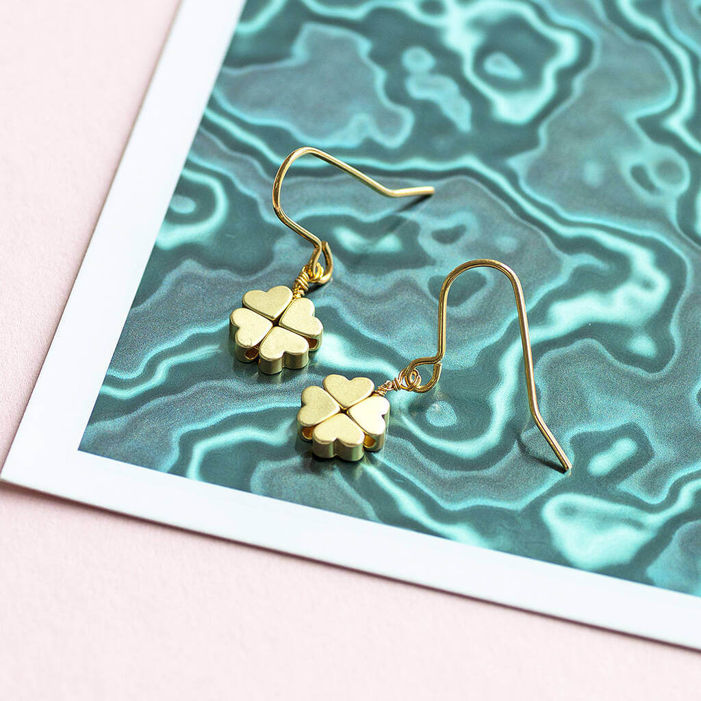 Gold plated four leaf clover earrings displayed on  a metallic green flat background.