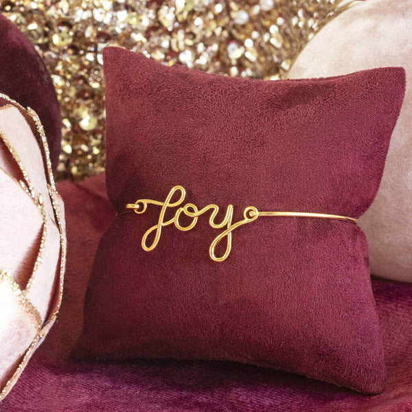 Image shows Gold Plated JOY Script Bracelet on a maroon jewellery pillow.