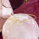 Image shows Gold Plated JOY Script Bracelet on a pink backdrop.