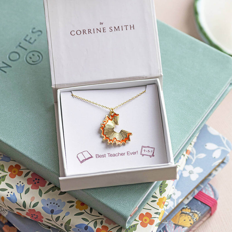 A gold plated necklace with pencil shaving charm trimmed in gold presented in a gift box on a Best Ever Teacher sentiment card set on top of a pile of notebooks.