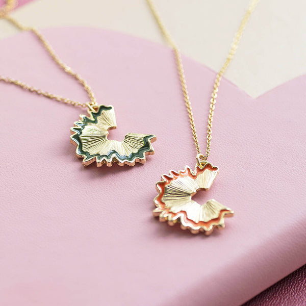 Two necklaces laid on top of a pink notebook. Both gold, the necklace on the left has a green trim and the necklace on the right has an orange trim.